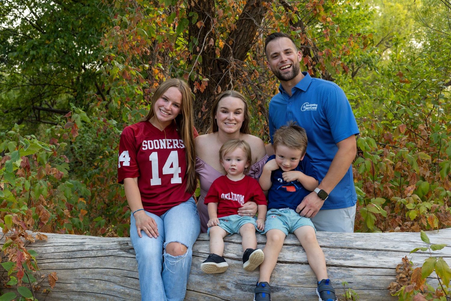Family of five smiling together on a log outdoors, wearing Sooners gear, surrounded by autumn foliage.