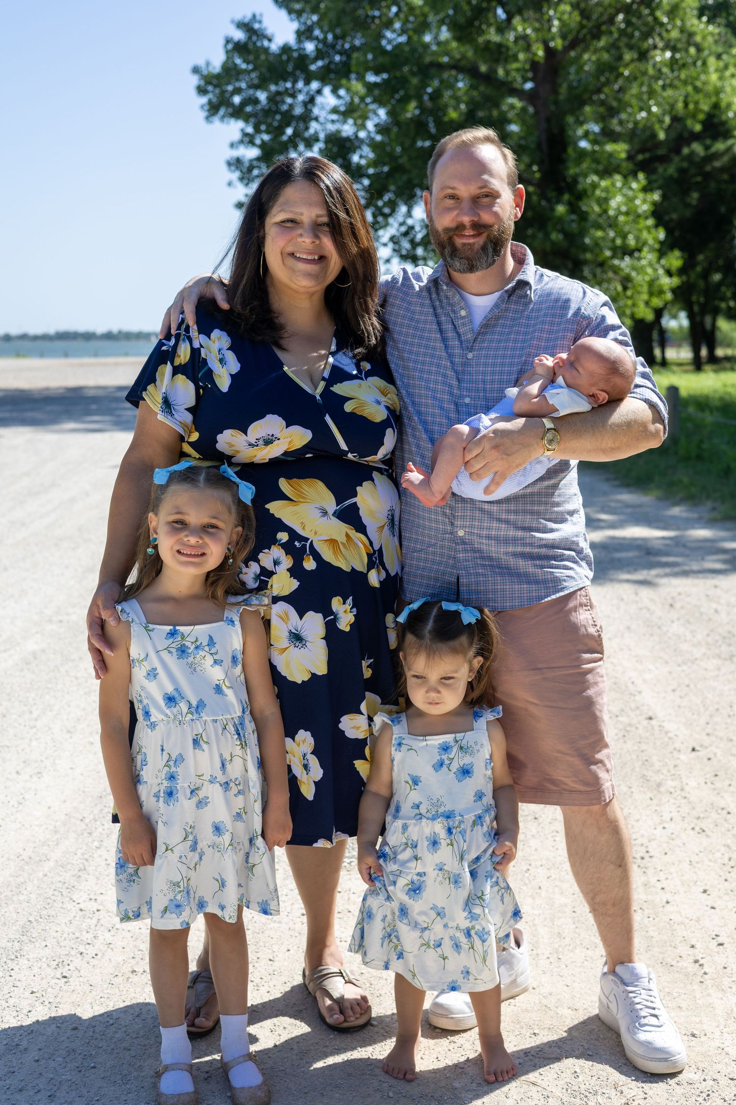Happy family of five posing outdoors near a beach, parents with three young children including a newborn, on a sunny day.