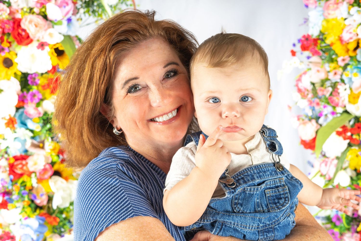 Smiling woman with red hair holds a cute baby in denim overalls in front of a colorful floral backdrop.