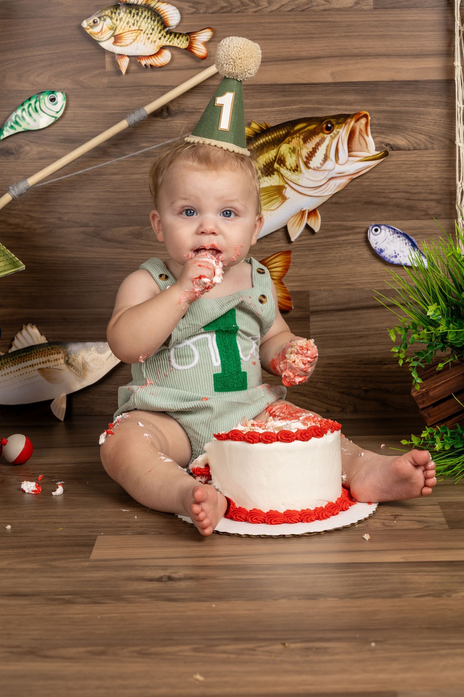 Baby boy in green romper smashing first birthday cake surrounded by fishing themed decorations on wooden floor.