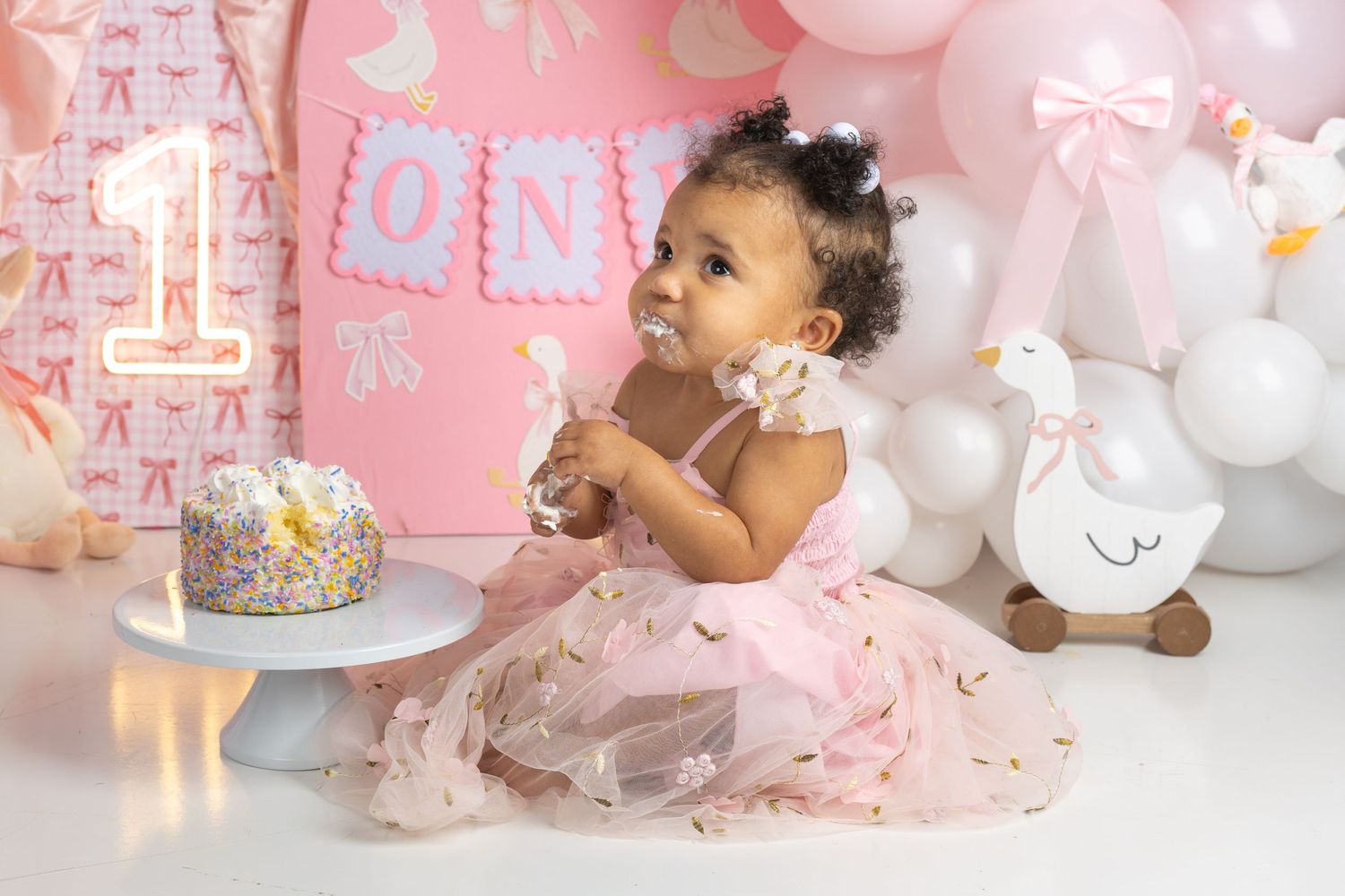 Baby girl in pink tutu dress smashing her first birthday cake surrounded by balloons and ONE decorations.