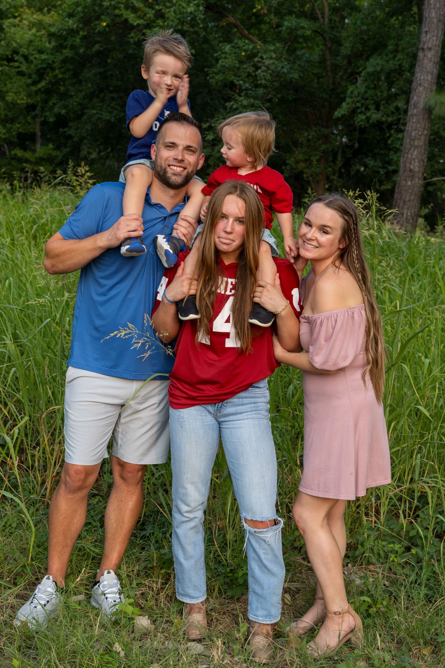 Happy family of five posing outdoors in green field, parents with two toddlers and a teen girl in sports jerseys.