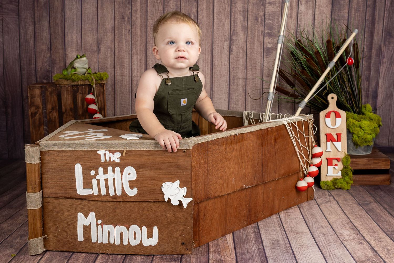 Baby boy in overalls sits in a wooden boat prop labeled The Little Minnow for a fishing-themed first birthday photo.