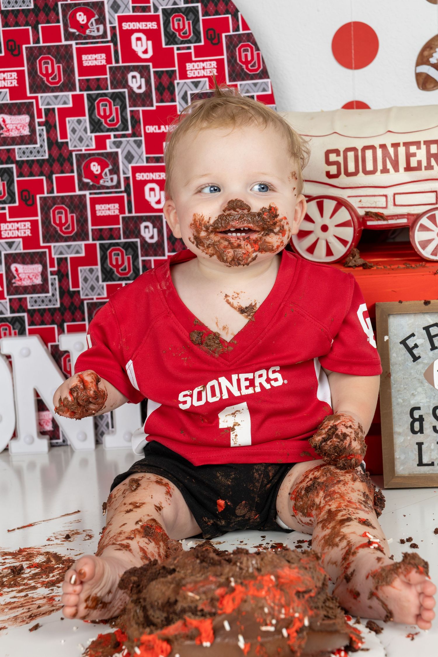 Baby in Sooners jersey covered in chocolate cake during first birthday smash cake photo session with OU themed backdrop.