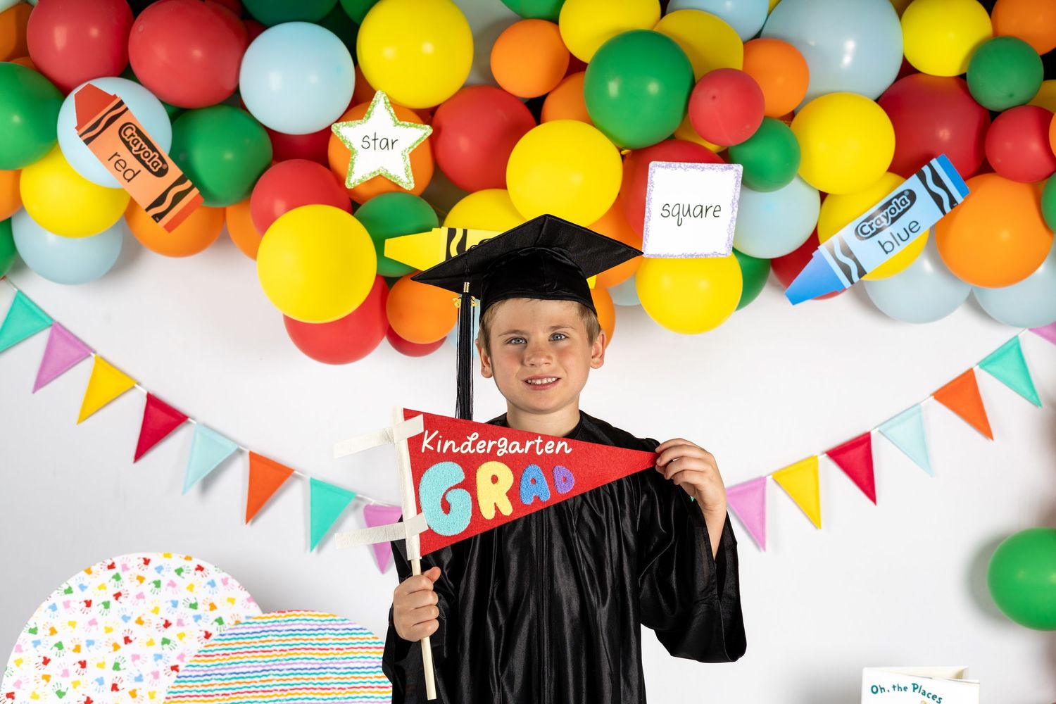 Young boy in graduation cap and gown holding a Kindergarten Grad banner, celebrating with colorful balloons.