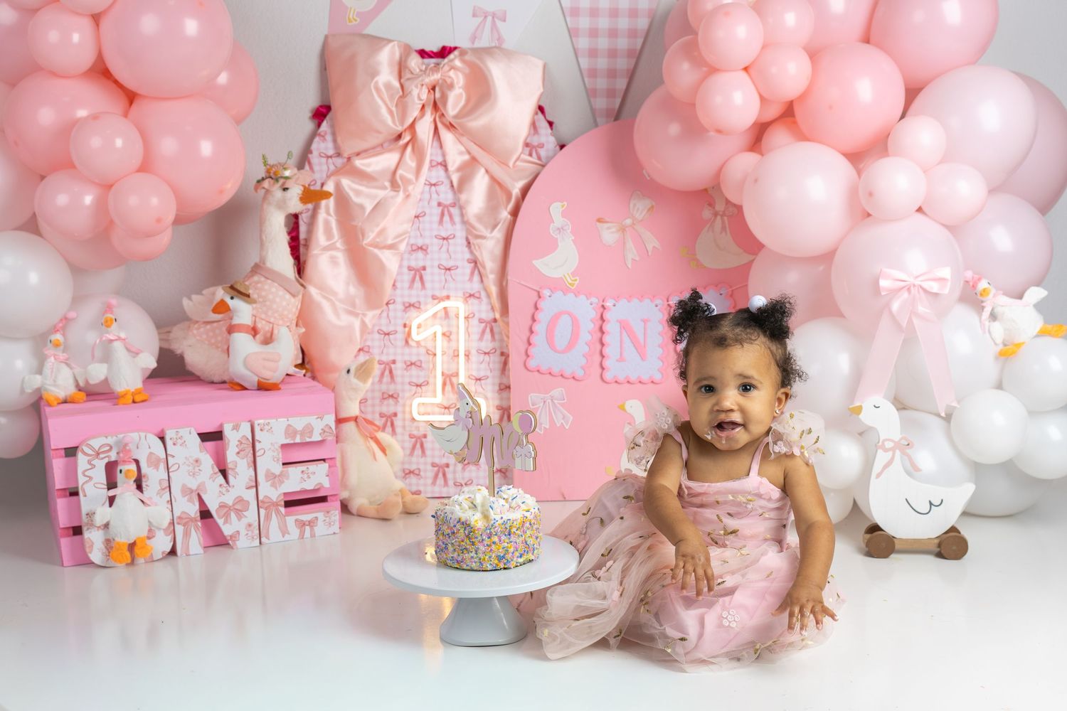 Baby girl in pink dress celebrates first birthday with cake, balloons, and ONE decorations in a pink party setup.