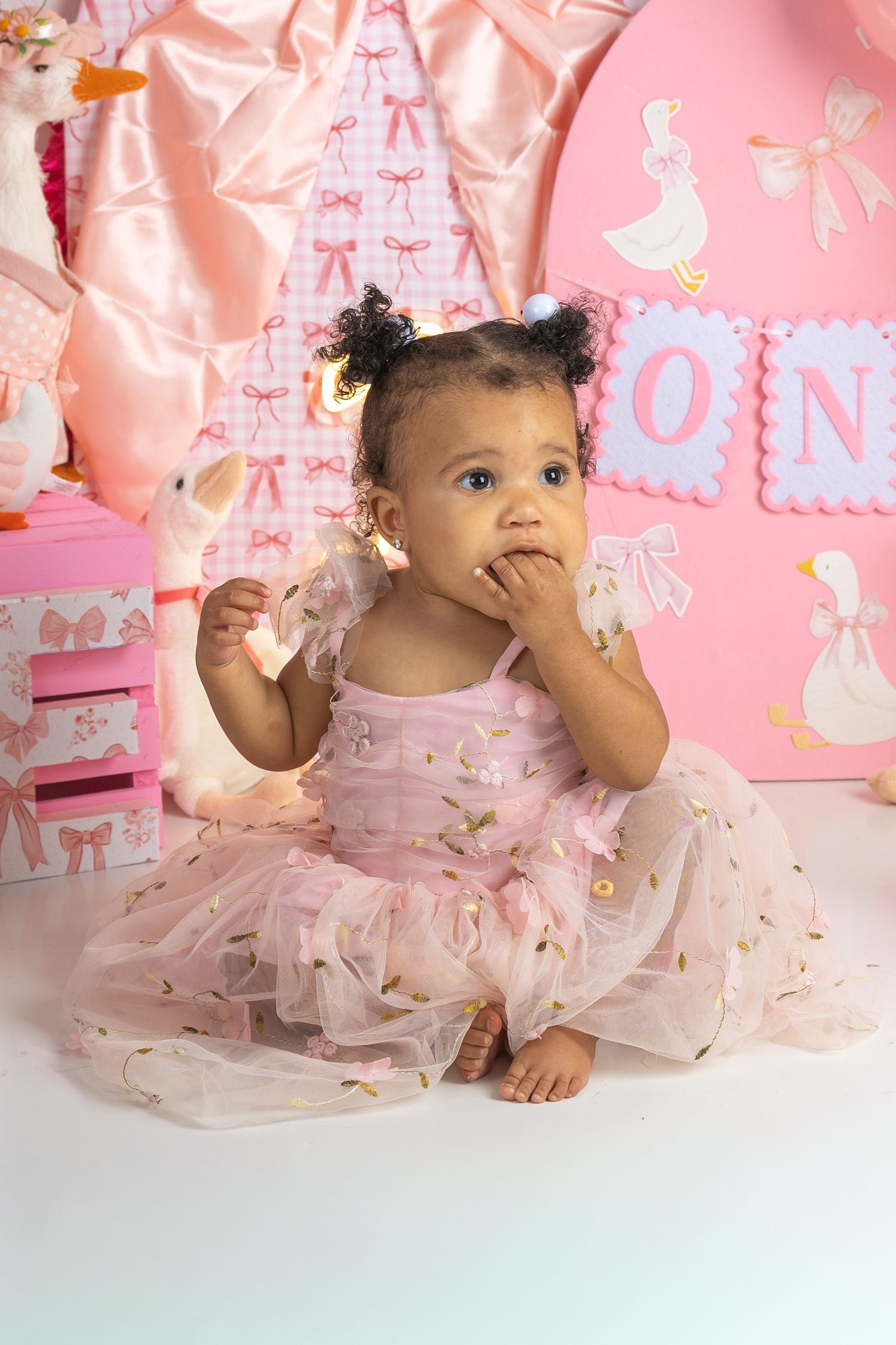 A baby girl in a pink tutu dress sits on the floor at a pink first birthday party setup.