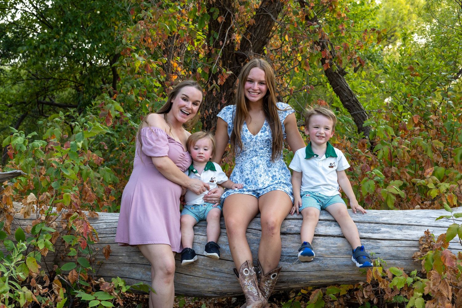 Family portrait of a woman with three children sitting on a log surrounded by lush green foliage outdoors.