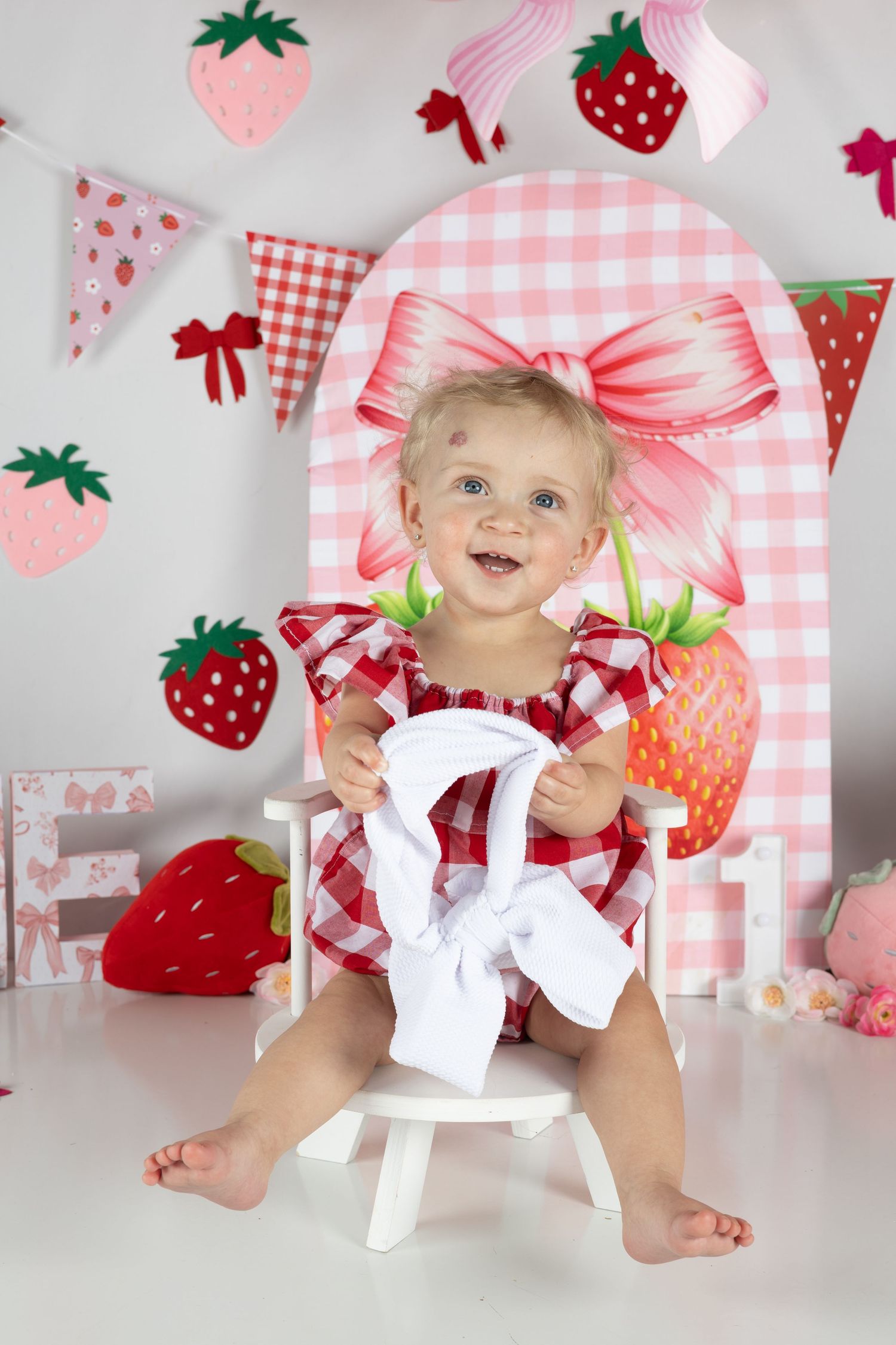 Smiling baby girl in red gingham dress sits in white chair with strawberry-themed party decorations backdrop.
