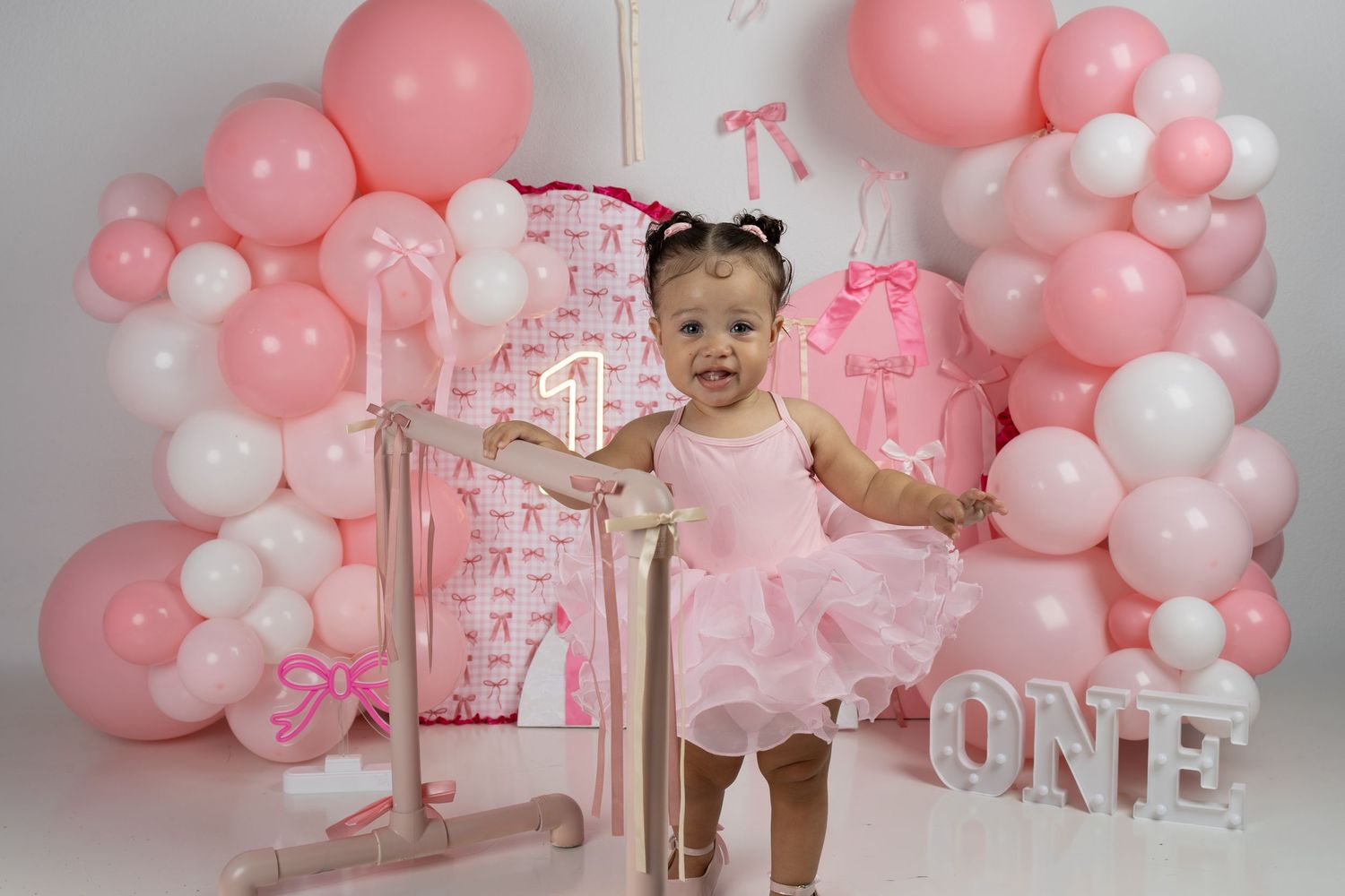 Baby girl in pink tutu celebrates first birthday surrounded by pink and white balloon garland with ONE sign.