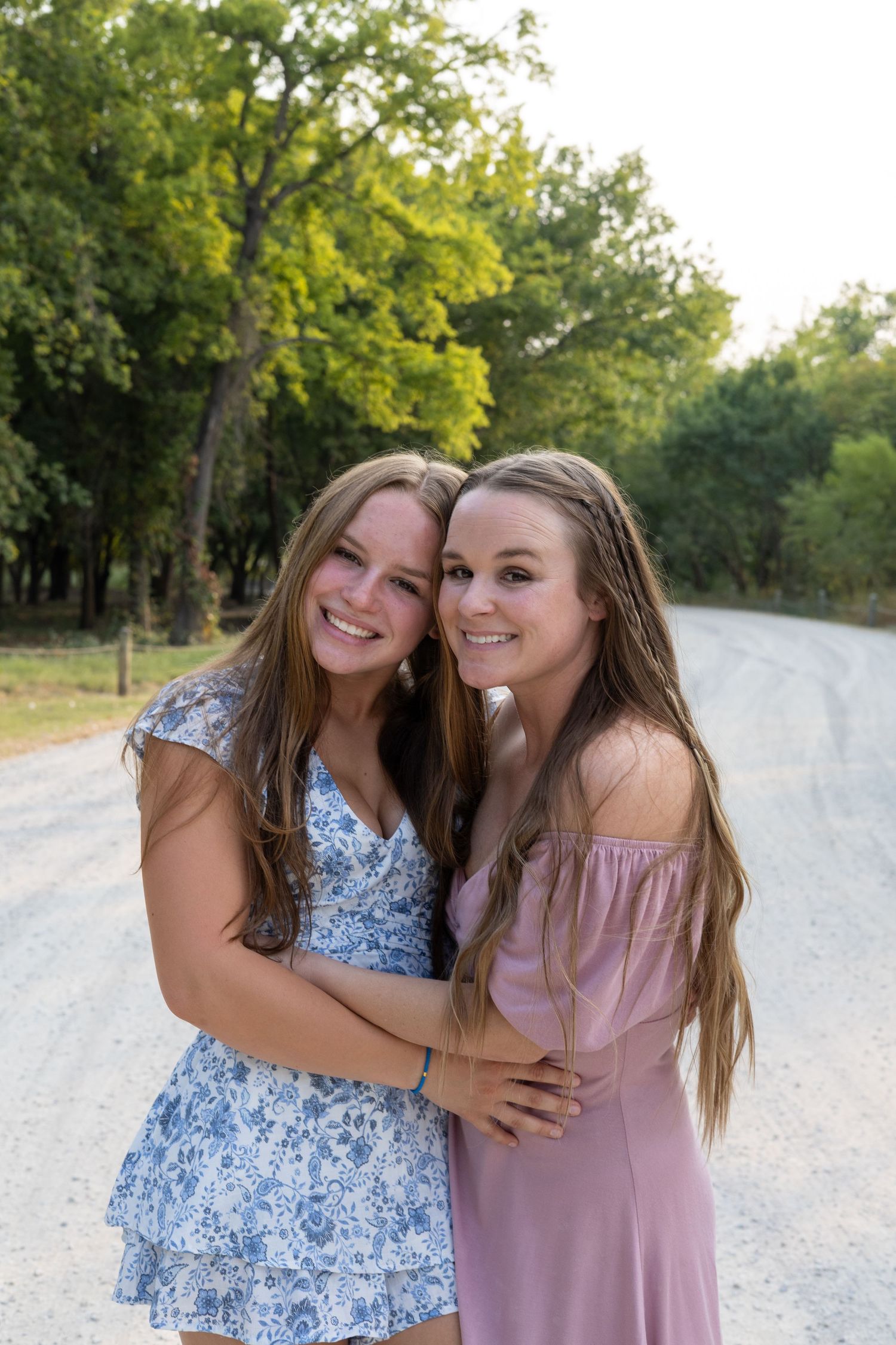 Two smiling teenage girls in summer dresses embrace on a rural dirt road surrounded by lush green trees.