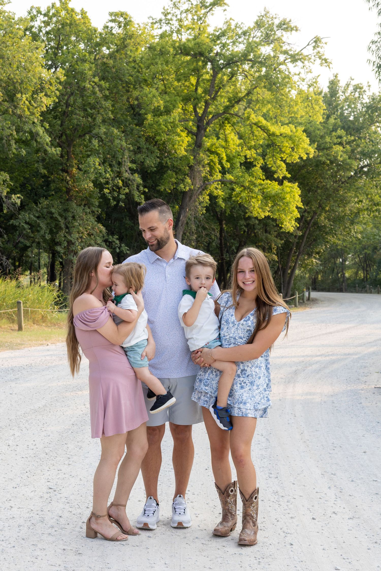 Family of five posing on a gravel path surrounded by green trees, with two adults holding toddlers and a teenage girl smiling.