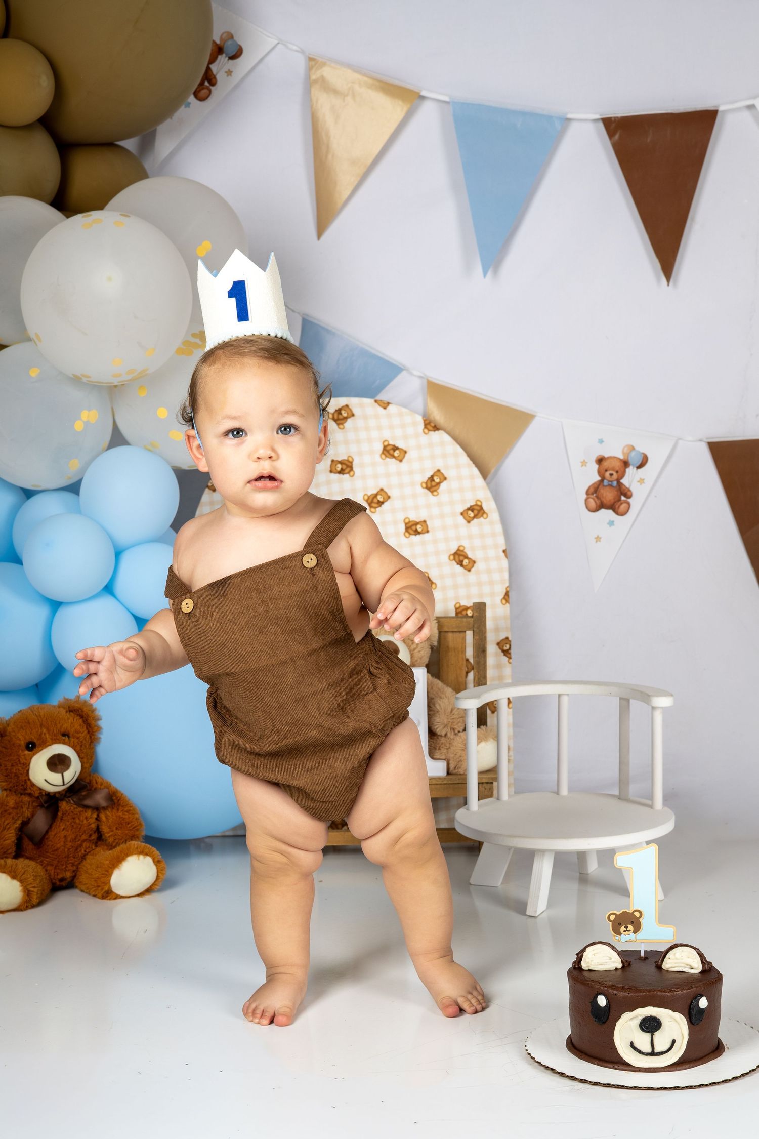 Baby's first birthday party with teddy bear cake, blue balloons, bunting, and crown in brown and blue themed setup.