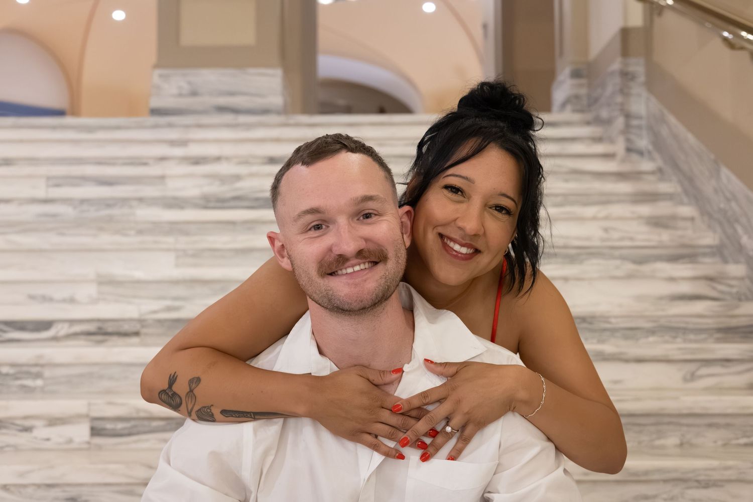 Happy couple posing on marble stairs, woman hugging man from behind, both smiling in white outfits.