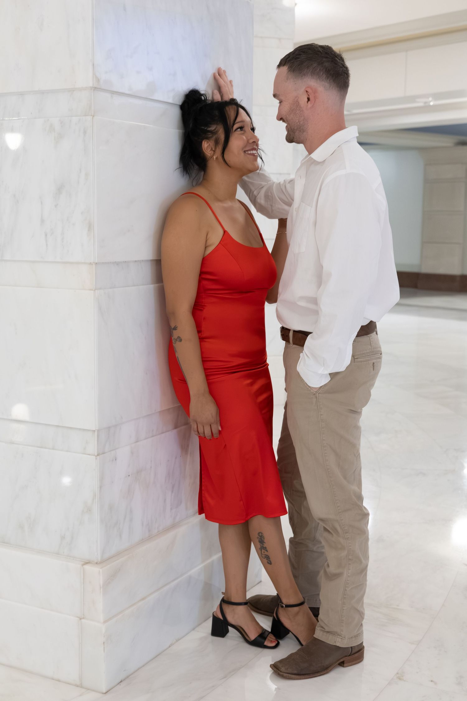 Couple posing romantically against a marble column, woman in red dress and man in white shirt and khaki pants.