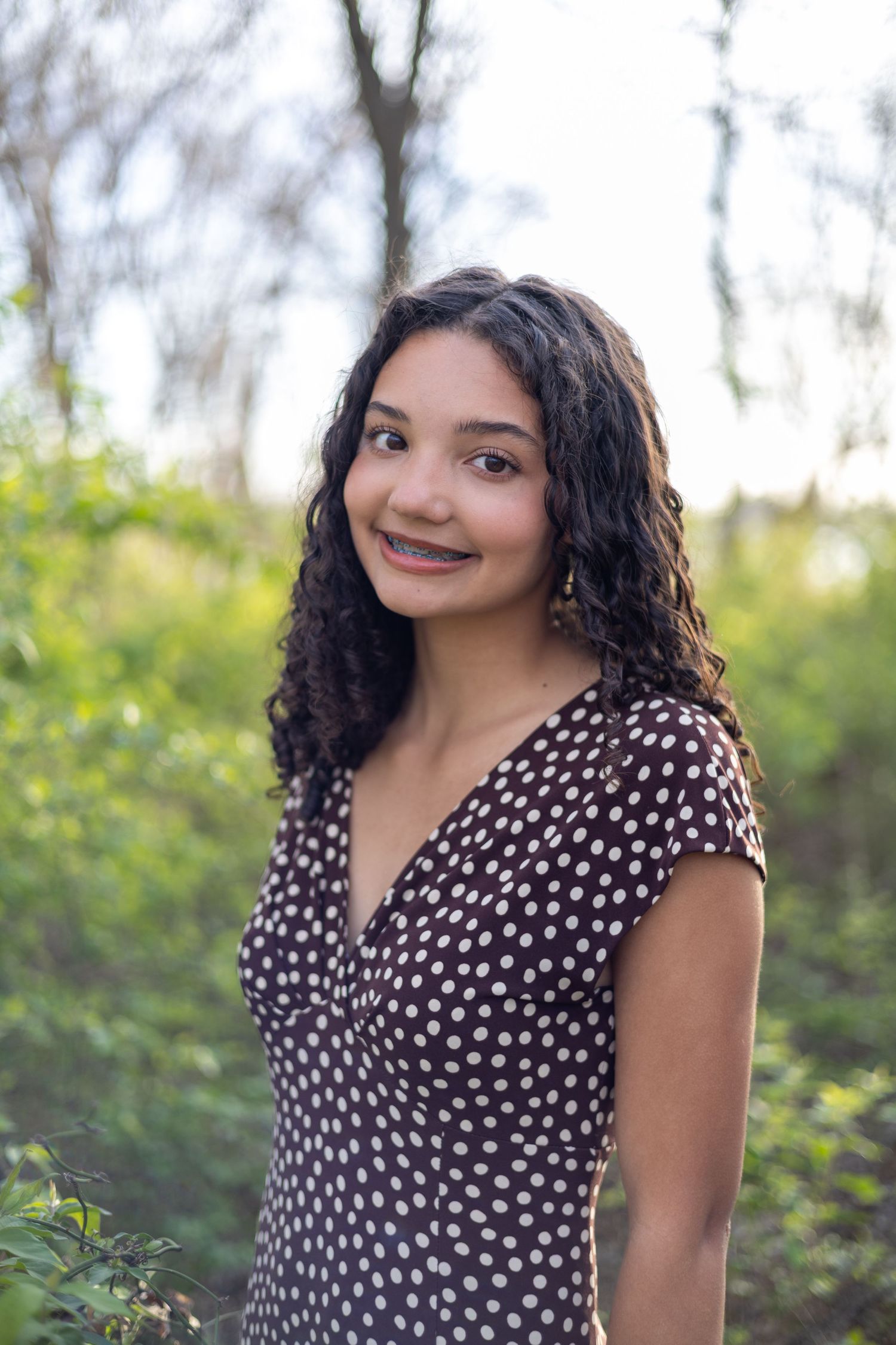 A person wearing a black and white polka dot dress poses outdoors in a natural setting with greenery in the background.