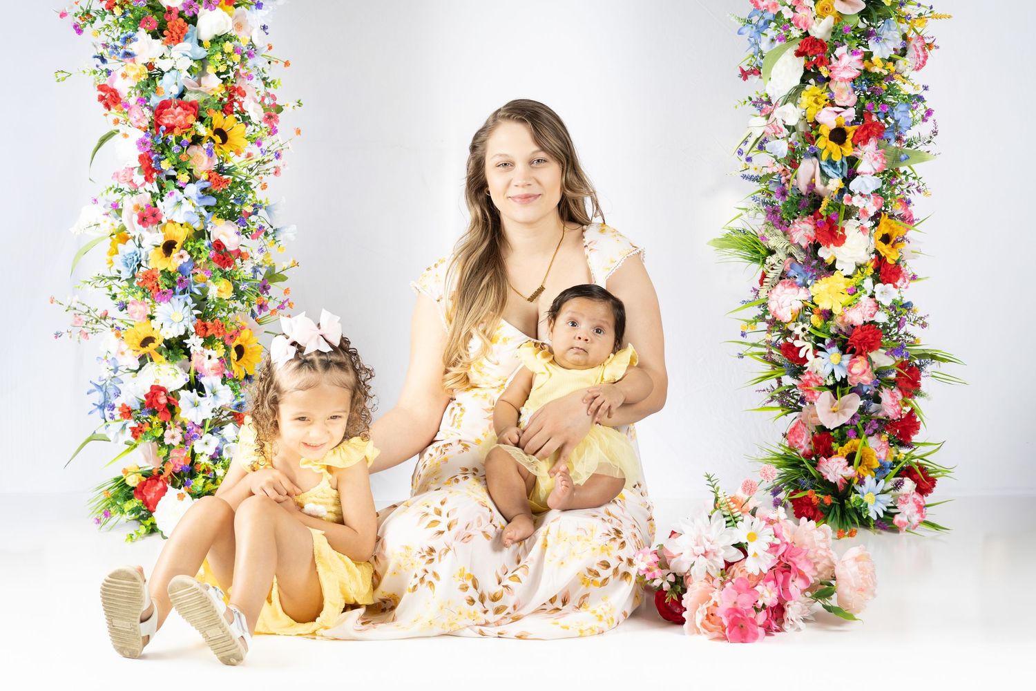 A mother in a yellow floral dress sits with two young children in yellow outfits against a white backdrop with colorful flower columns.