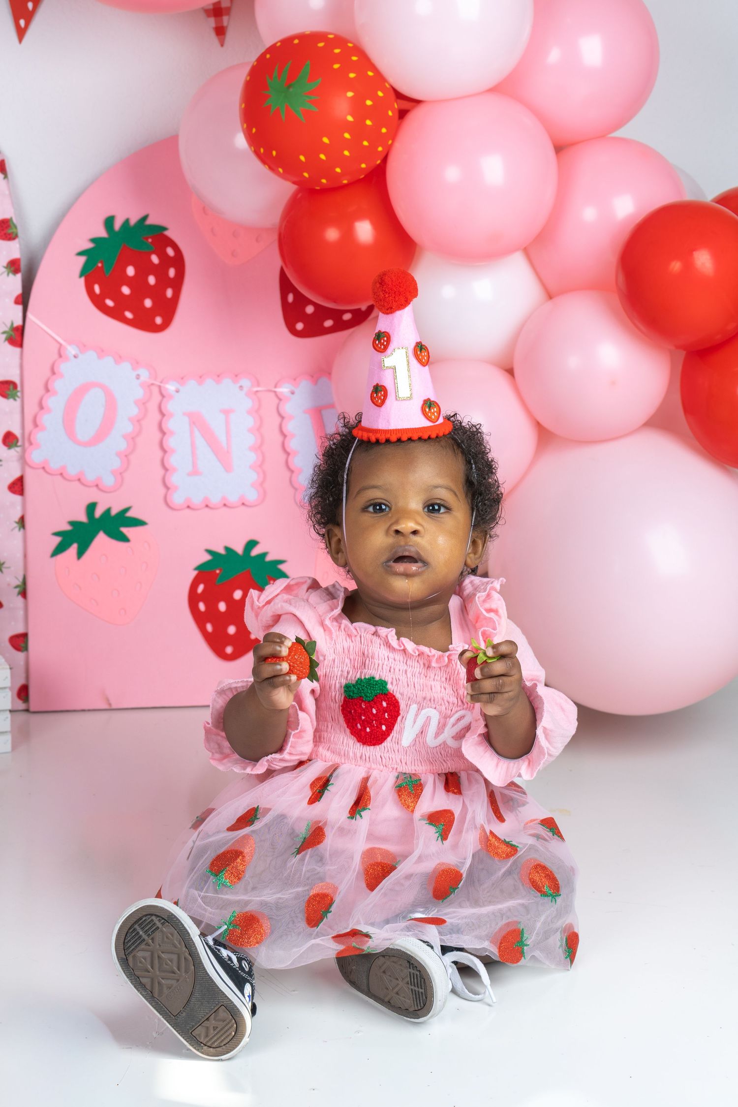 Baby girl in strawberry dress and party hat celebrates first birthday with pink and red balloon decorations.