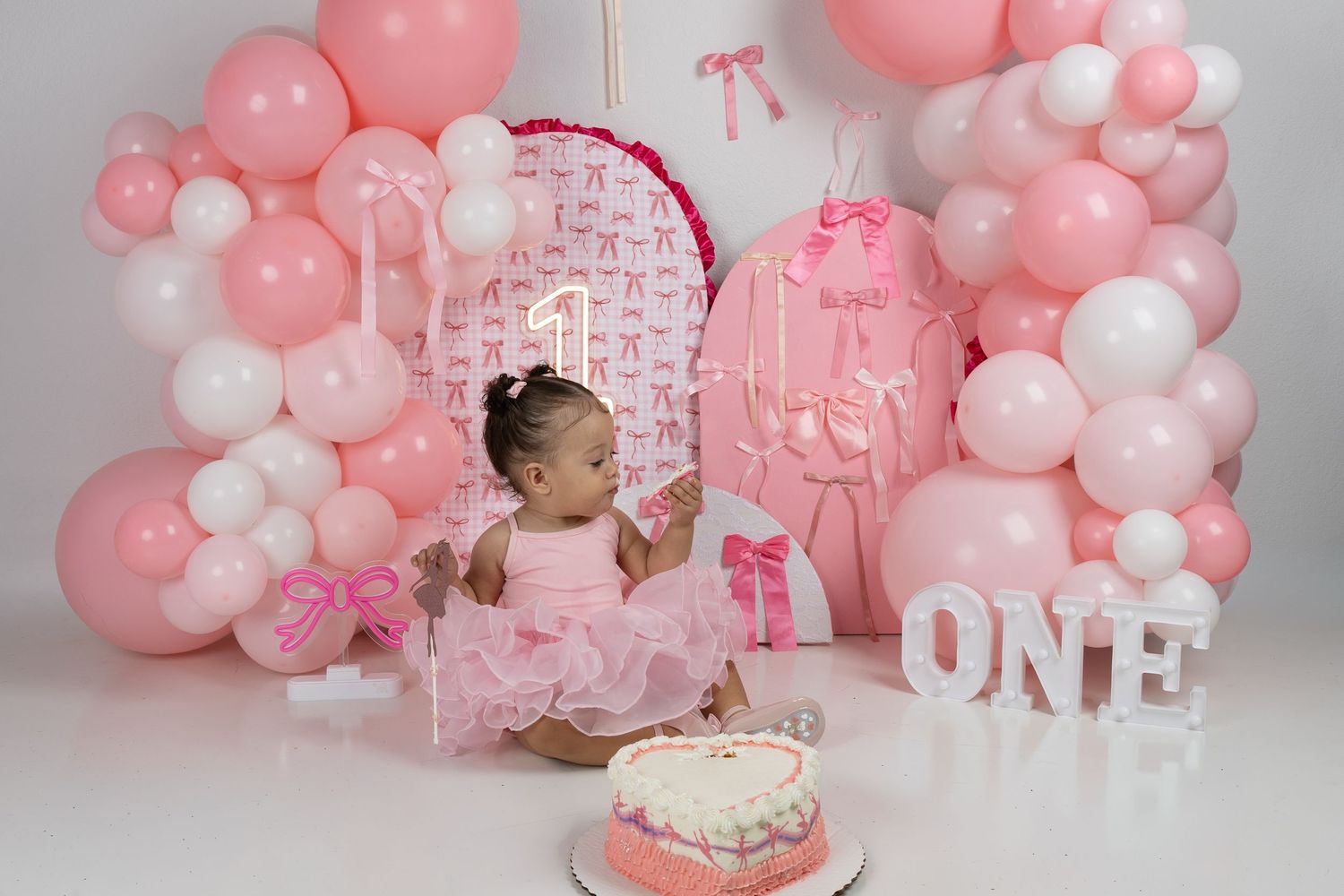 Baby girl in pink tutu sits before a first birthday cake with pink balloon arch and ONE sign decorations.