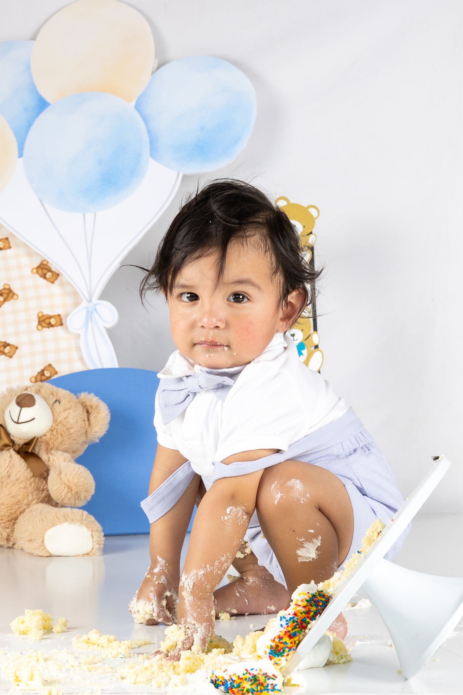 Baby's first birthday cake smash with teddy bear, balloons, and colorful sprinkles on white background.