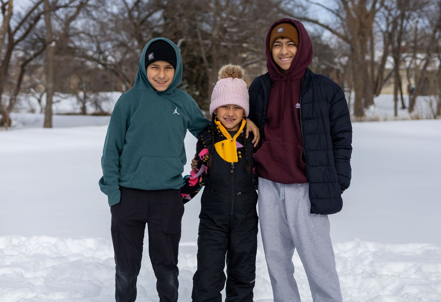 Three people wearing winter clothing stand together in a snowy park on a cold day.