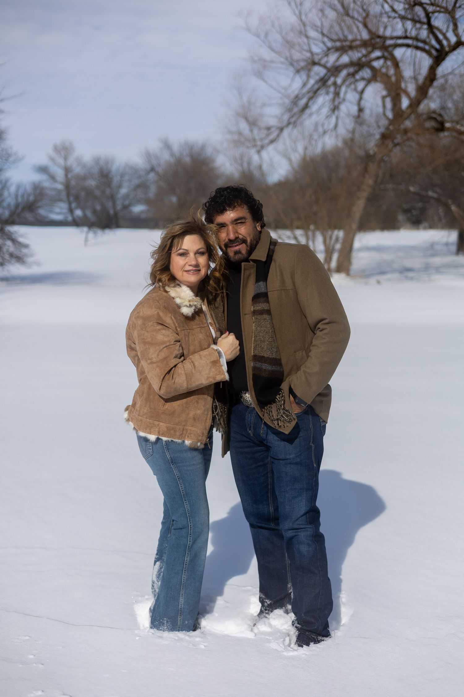 A couple stands together in a snowy winter landscape wearing warm coats and jeans.