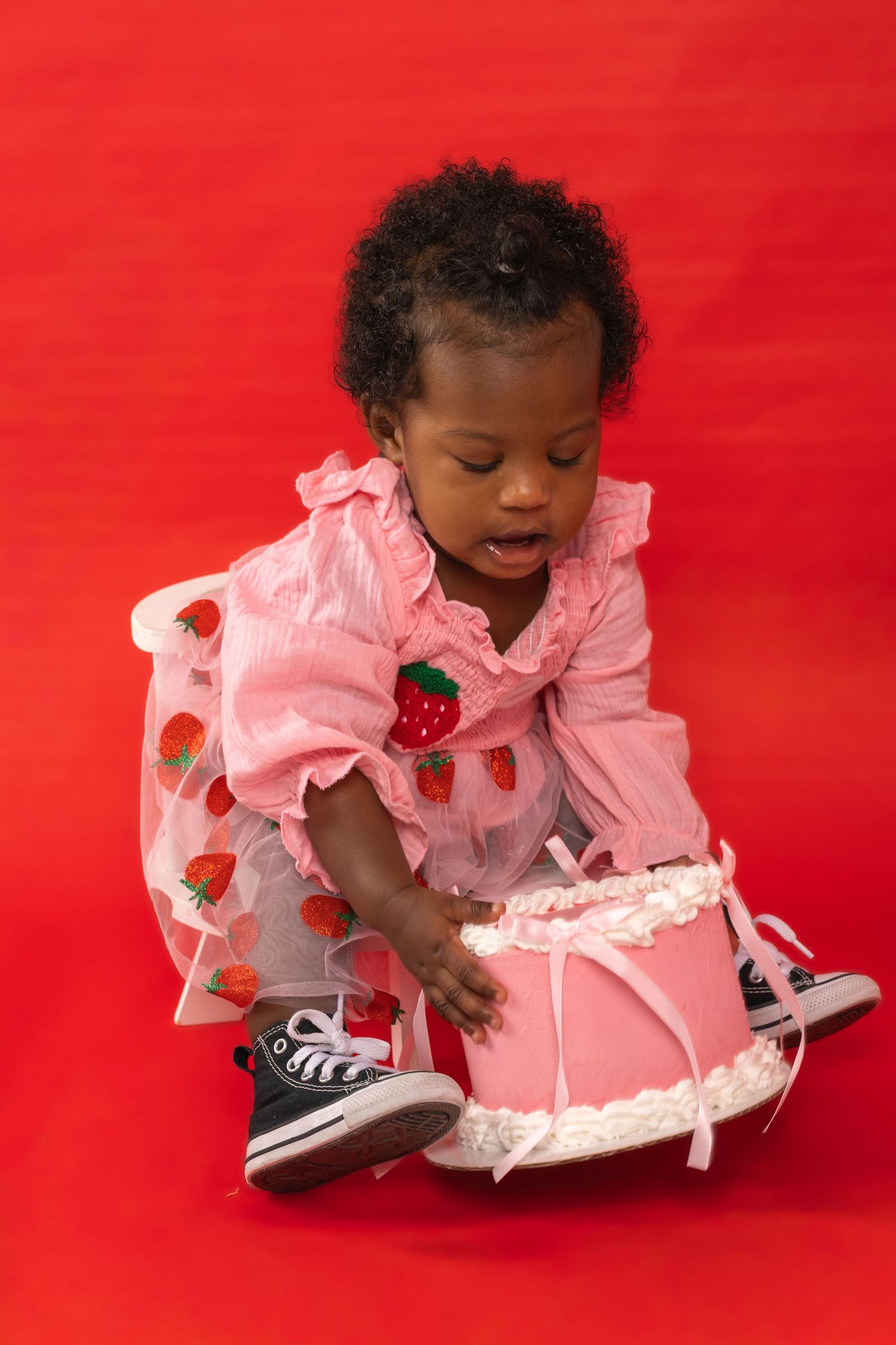 Toddler girl in pink strawberry dress and Converse sneakers reaching for a pink birthday cake on red background.