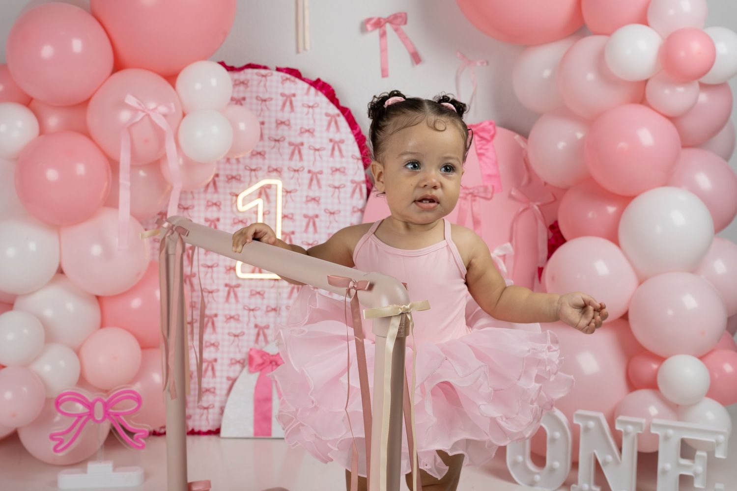 Baby girl in pink tutu poses at her first birthday party with pink balloon arch and number one decorations.
