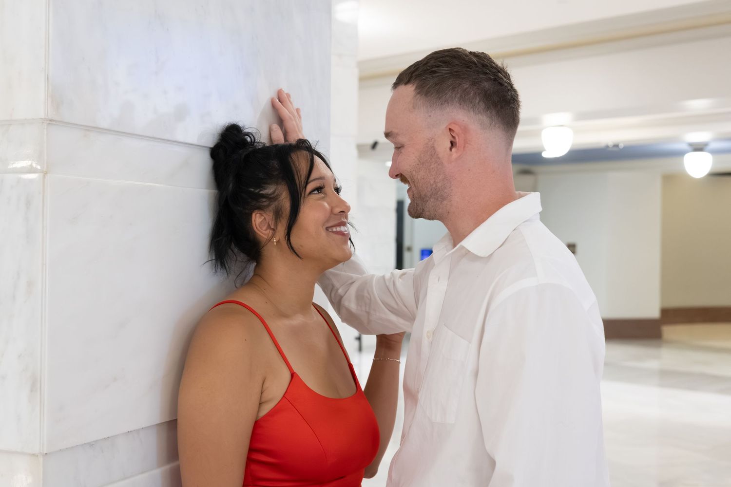 Smiling couple sharing a romantic moment leaning against a marble wall, woman in red dress, man in white shirt.