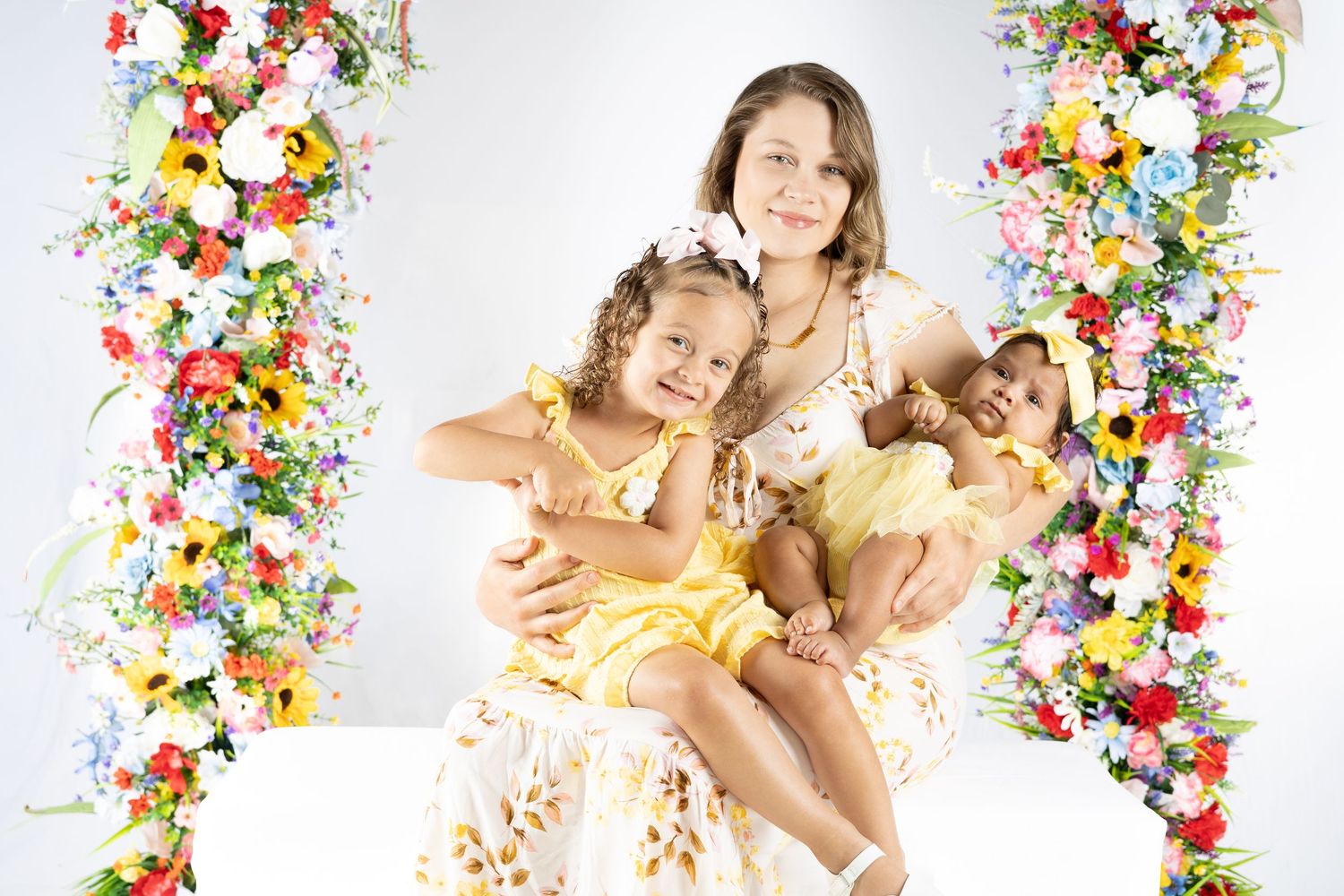 A mother in floral dress holds two young girls in yellow dresses on a swing adorned with colorful flowers.