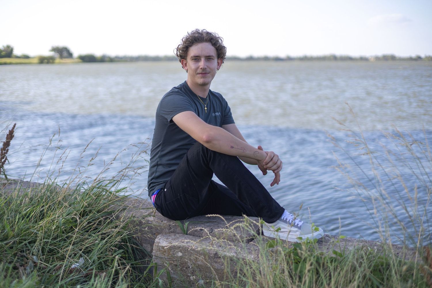 Young man sitting on a stone ledge by a calm lake, wearing a black t-shirt and jeans, with green grass nearby.