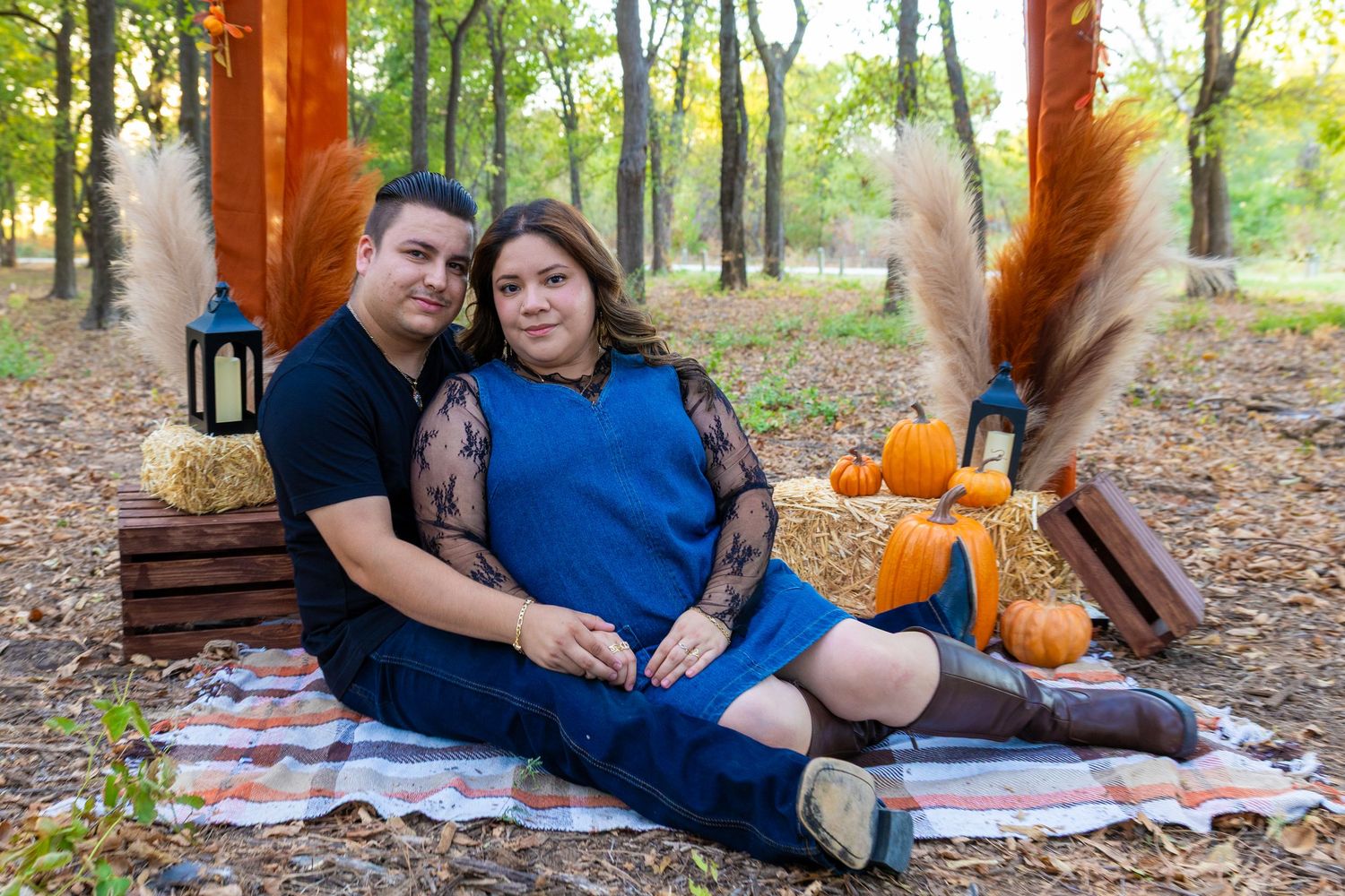 Couple sitting on a plaid blanket surrounded by pumpkins and hay bales during a fall outdoor maternity photo session.