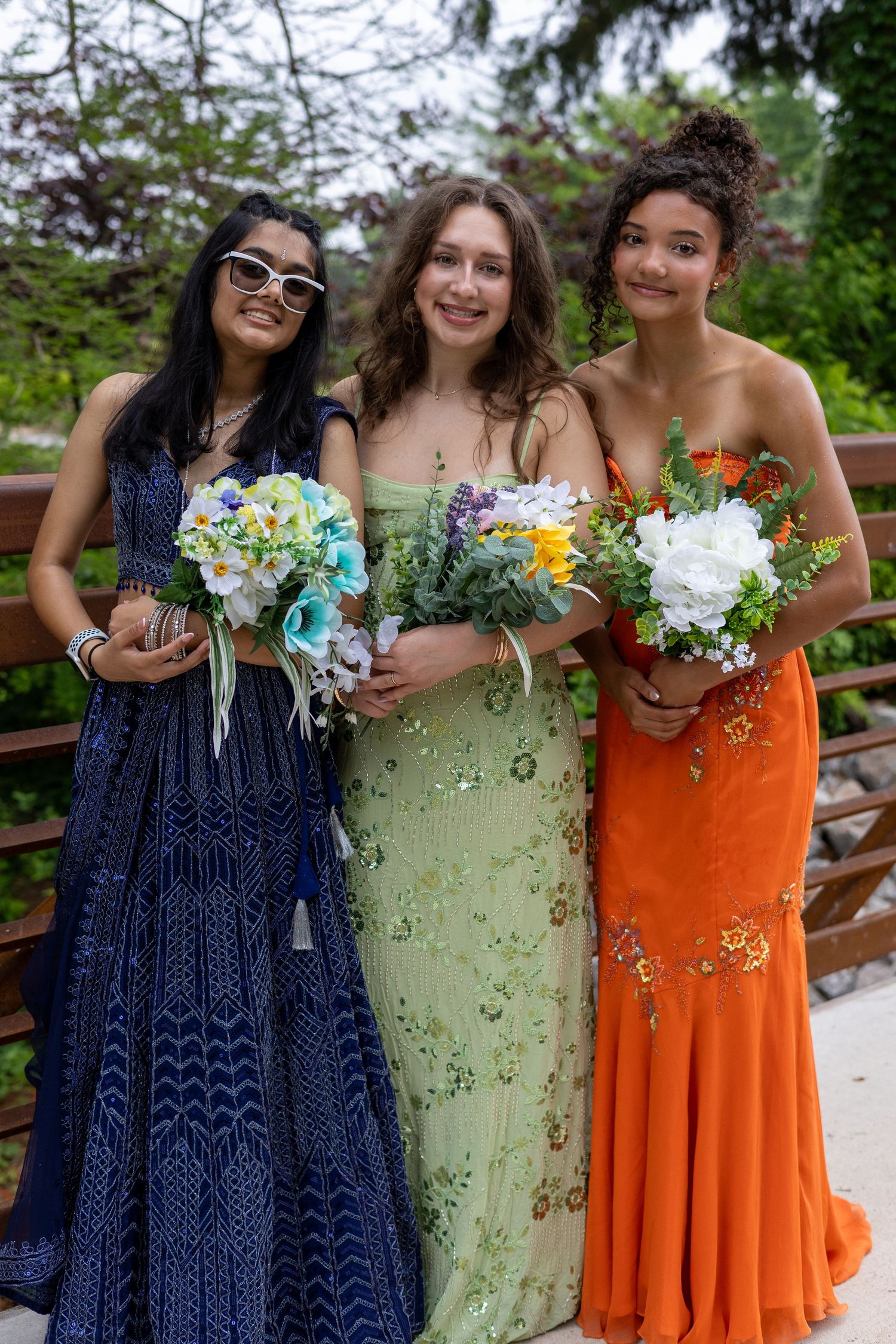 Three young women in elegant prom dresses holding bouquets, posing outdoors on a bridge surrounded by greenery.