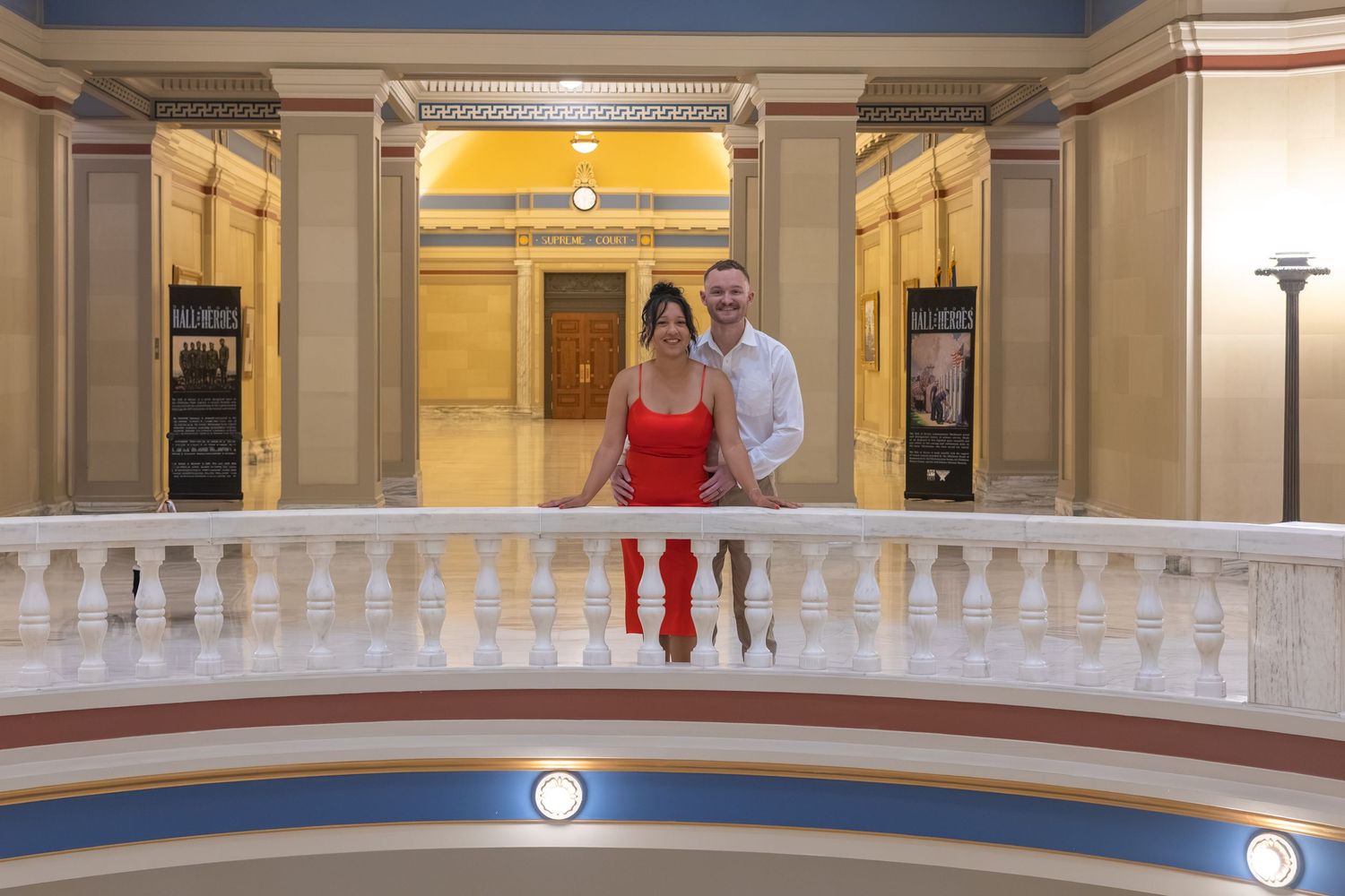 Couple posing on ornate white balcony in elegant neoclassical building with columns and yellow walls.