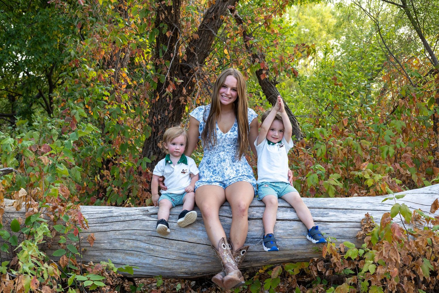 A smiling woman sits on a fallen log with two young toddlers in a lush green autumn forest setting.