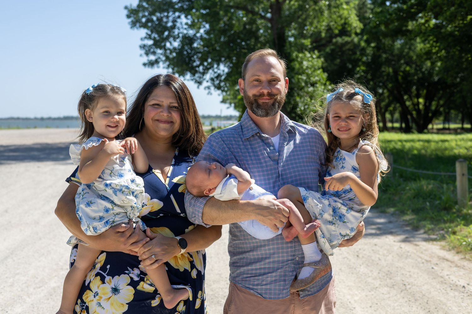 Happy family of five posing outdoors on a sunny day, parents holding three young children on a rural gravel road.