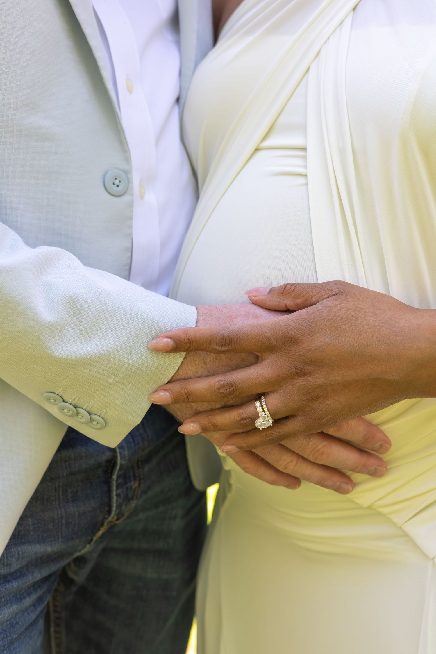 Pregnant couple holding baby bump, wearing wedding rings, man in white blazer and jeans, woman in yellow dress.