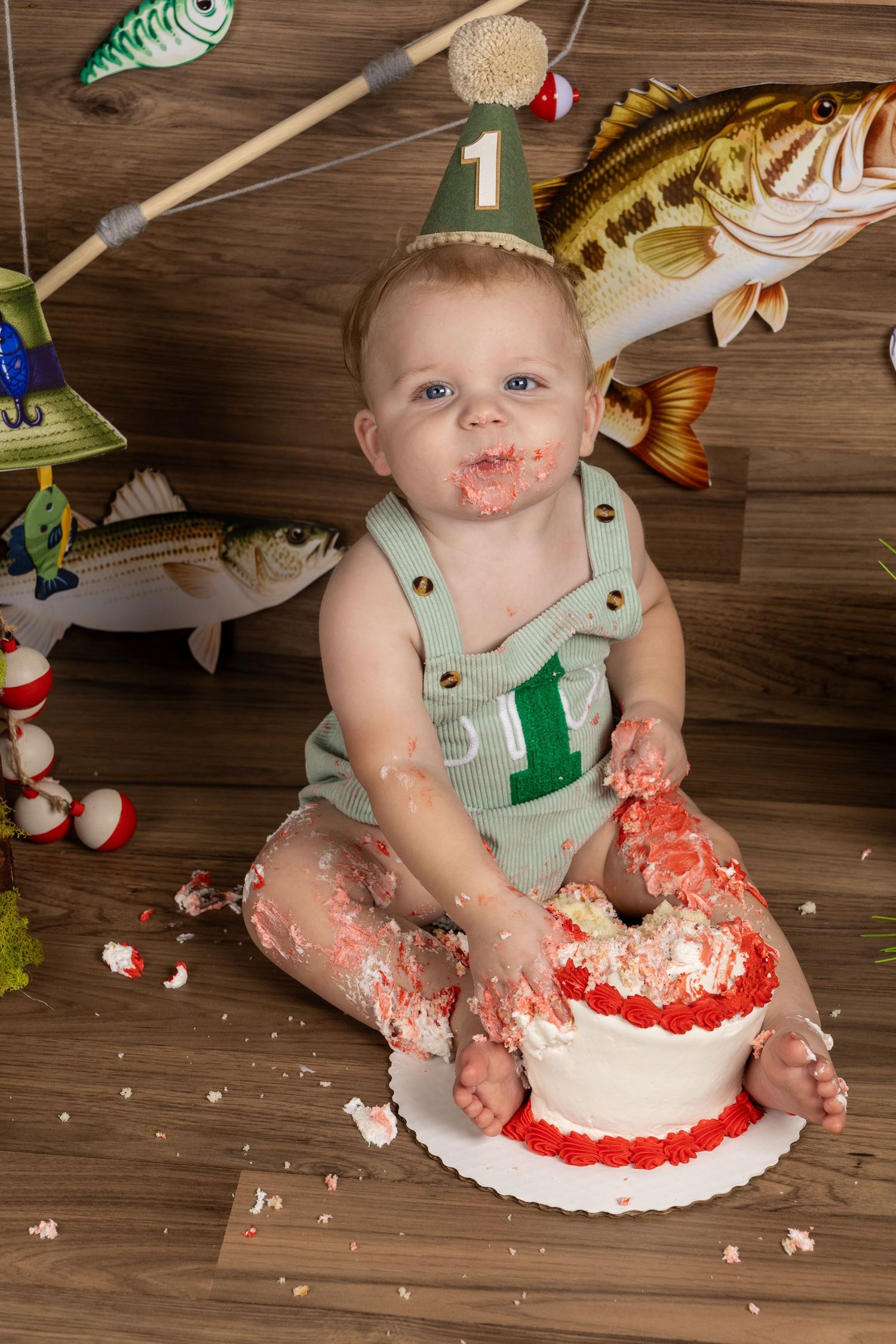 Baby boy in overalls smashes a white and red cake during a fishing-themed first birthday cake smash photo session.