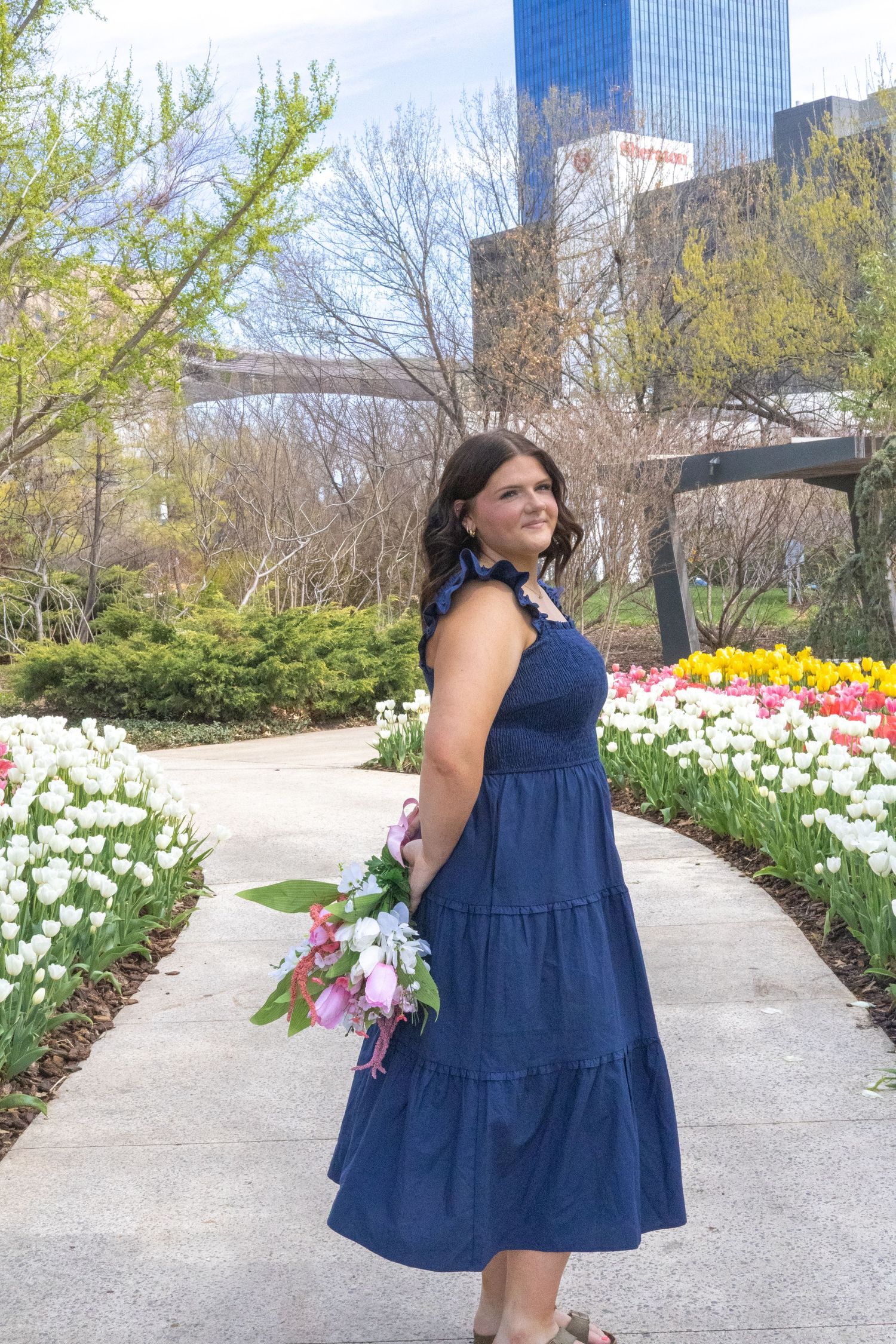 A garden path winds through blooming spring tulips and flowers near modern buildings on a sunny day.