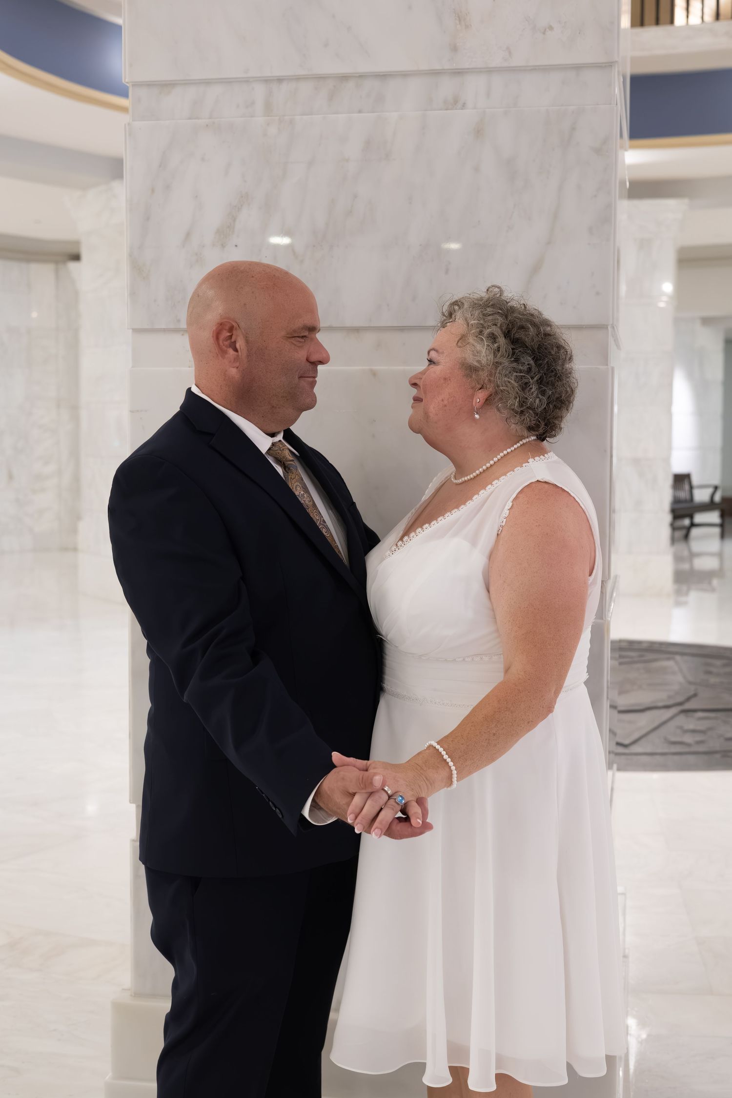 Older couple in wedding attire holding hands and smiling at each other in an elegant marble hall.