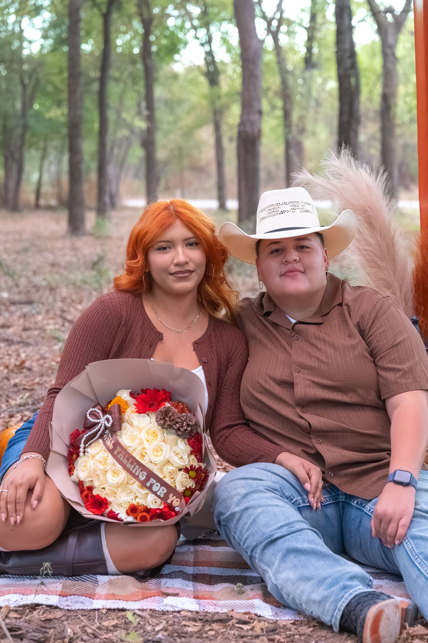 Couple sitting outdoors in woods, woman with red hair holding bouquet, man in white cowboy hat, on plaid blanket.