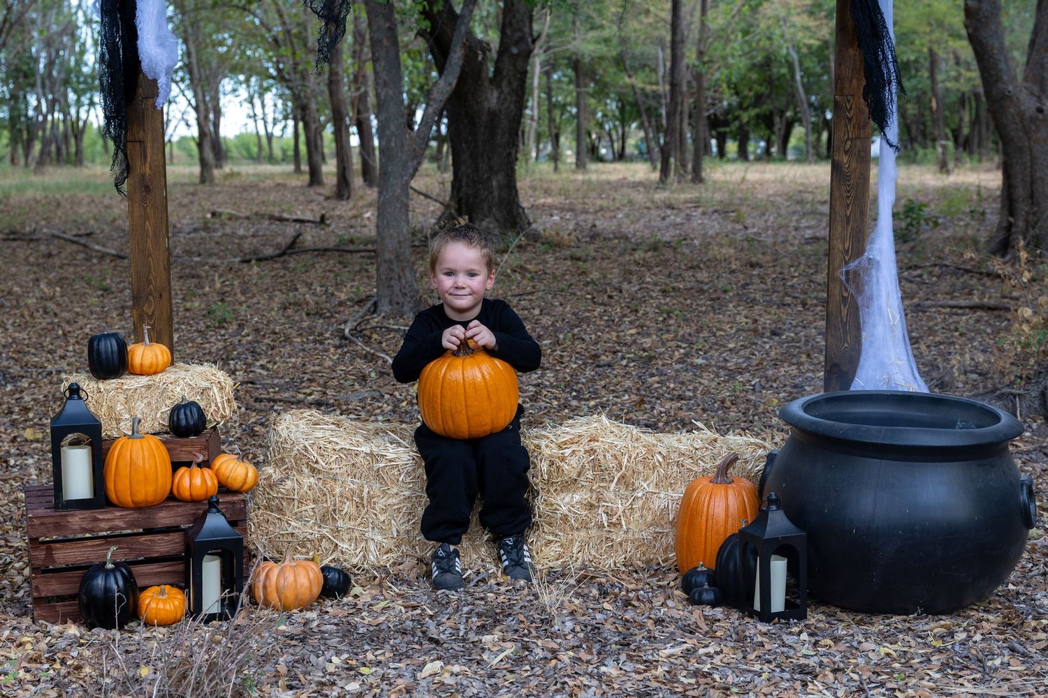 Child in black Halloween costume holding pumpkin on hay bale surrounded by pumpkins and spooky forest decorations.