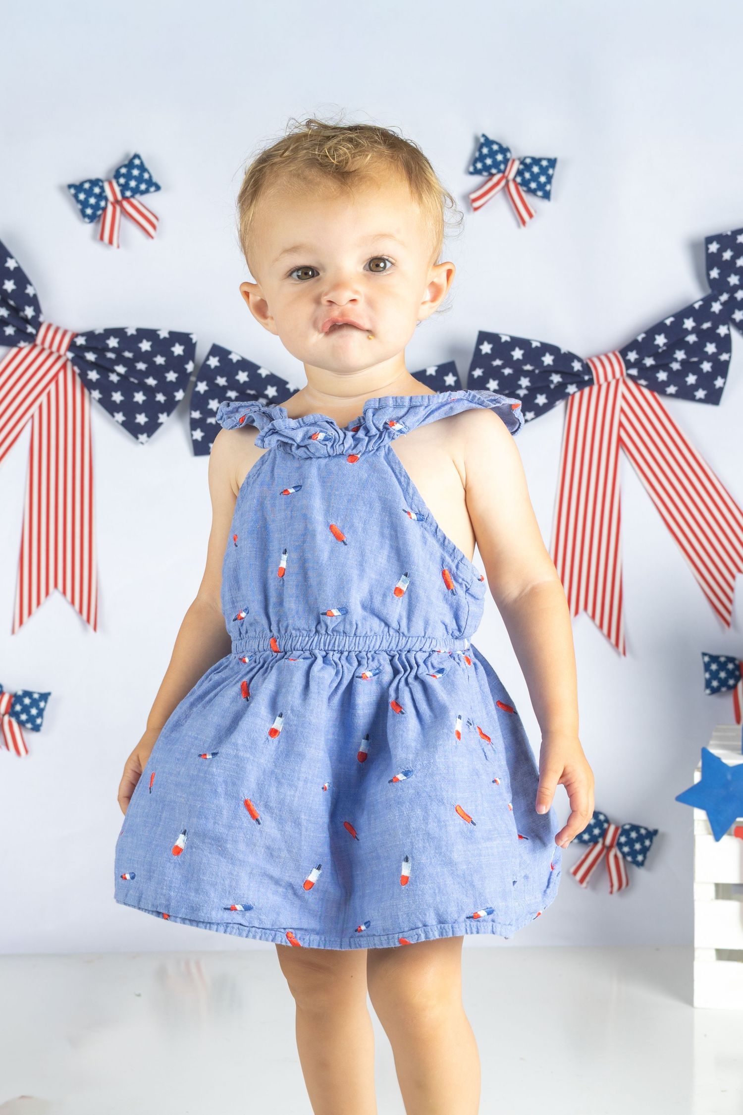 Baby girl in blue patterned dress poses in front of patriotic American flag decorations and bows.
