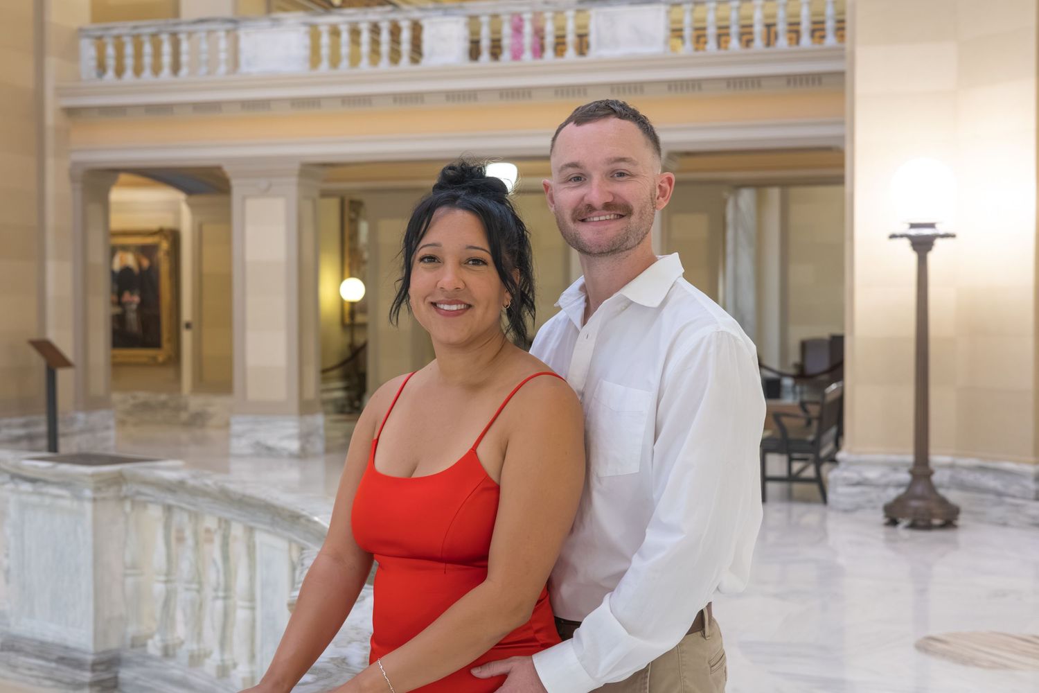 A smiling couple poses together in an elegant marble building interior, woman in red dress, man in white shirt.