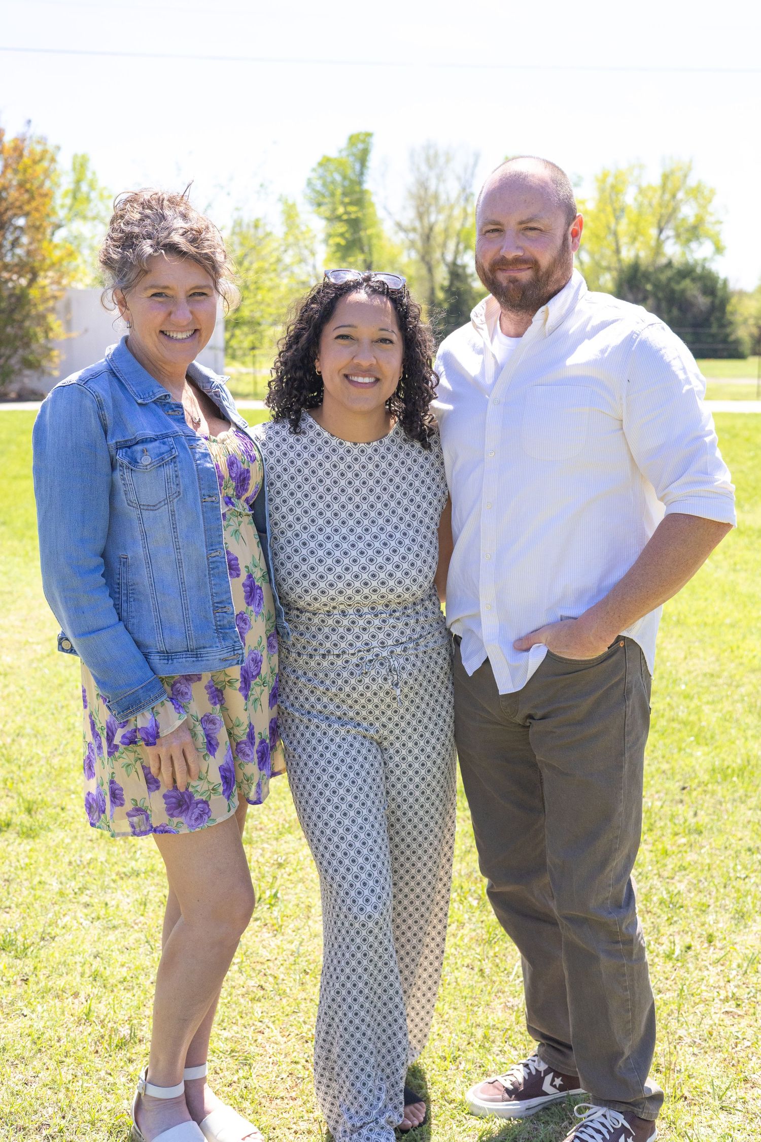 Three smiling friends pose together outdoors on a sunny day in a green park.