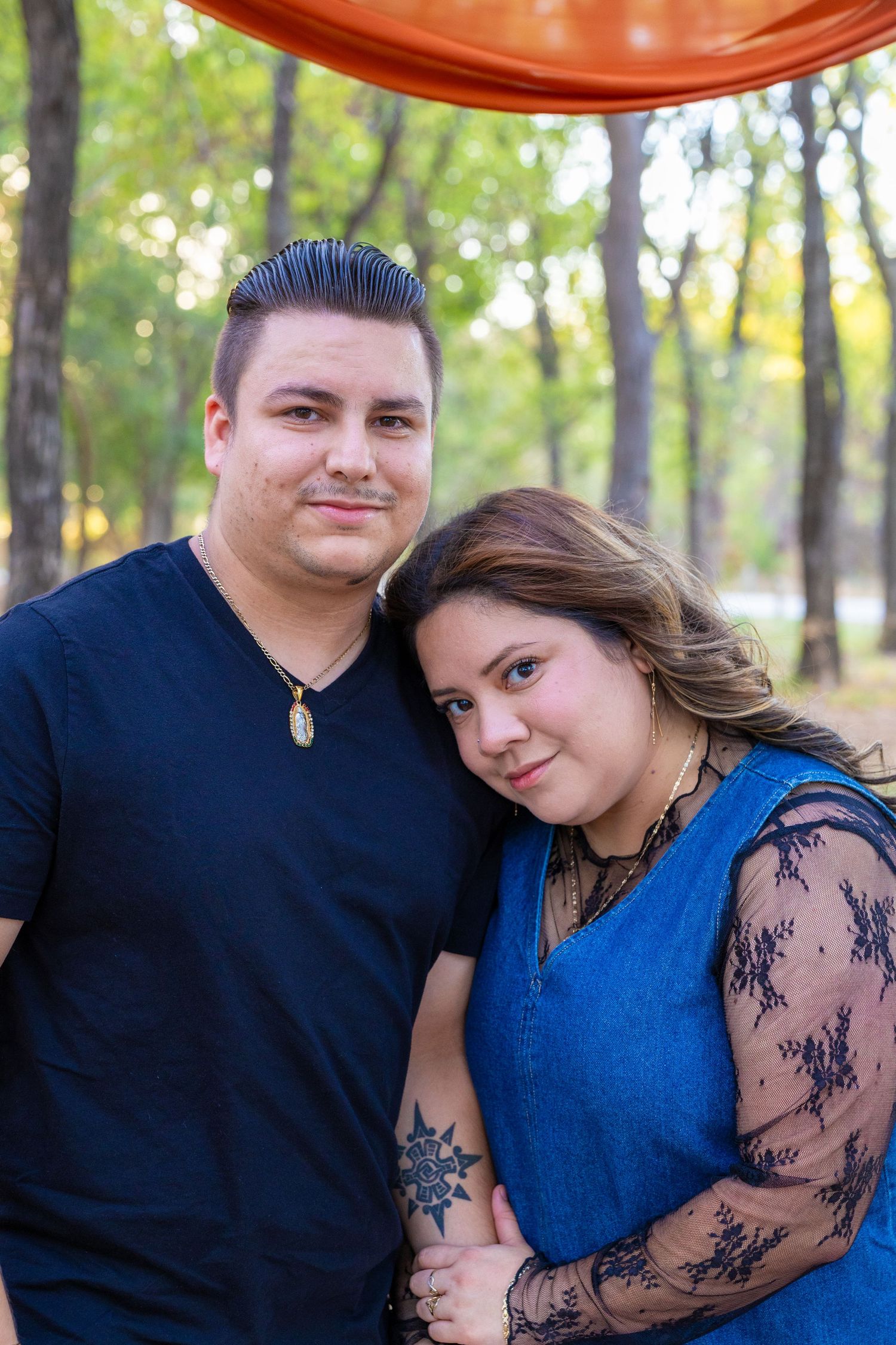 A couple poses outdoors in a wooded park, the woman leaning on the man's shoulder, both smiling warmly.