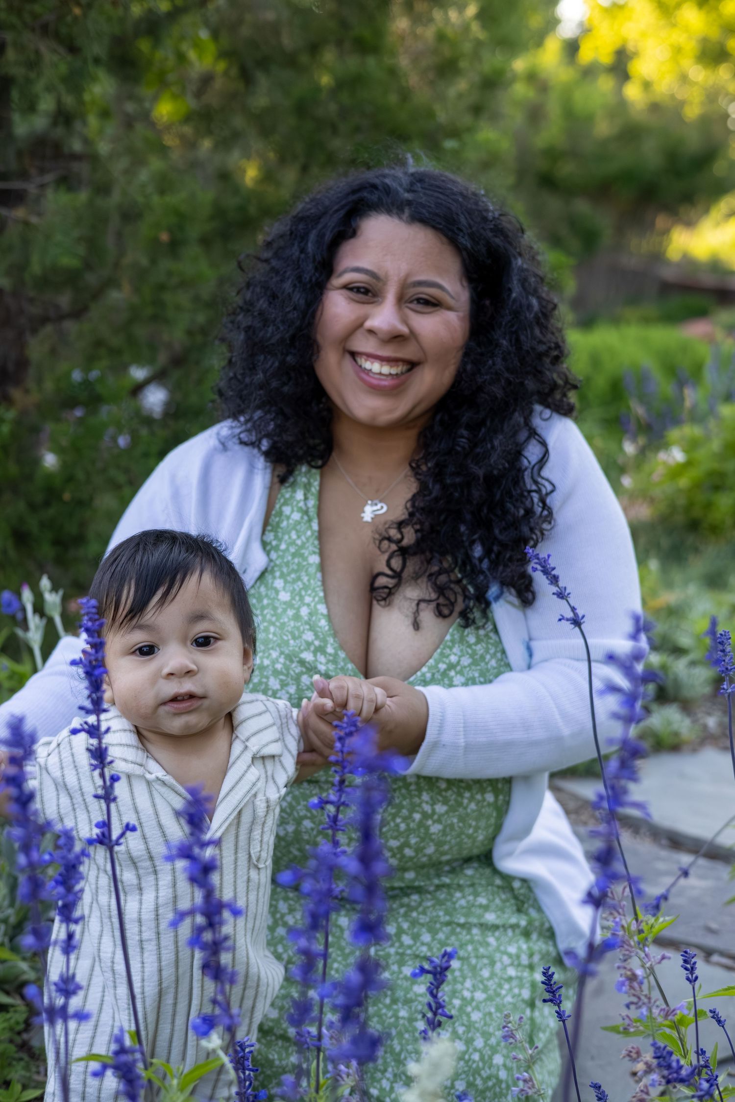 Smiling woman with curly hair holds baby boy among purple salvia flowers in a lush garden setting.