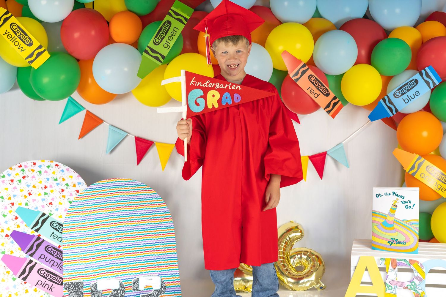 Child in red graduation cap and gown holding Kindergarten Grad sign, surrounded by colorful balloons and crayon decorations.