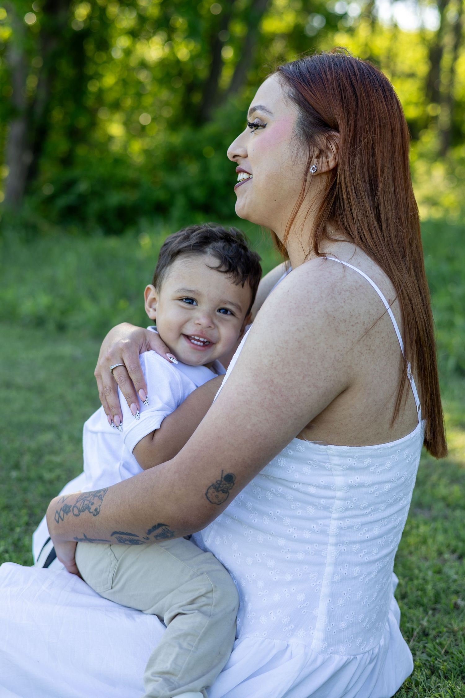 A smiling mother in a white dress holds her happy toddler on her lap outdoors in a lush green park.