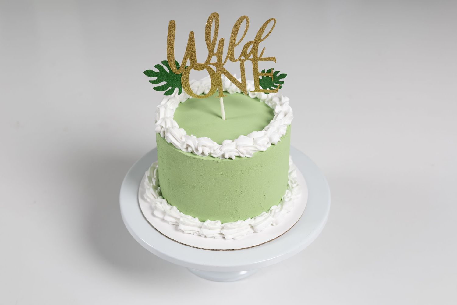Green frosted Wild One first birthday cake with white ruffles and gold topper on white cake stand.